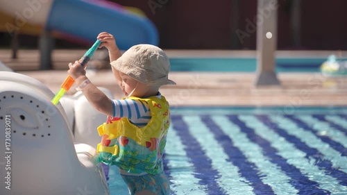 A happy little boy is enjoying himself in the pool. He is playing with a water gun and shooting at his younger brother.