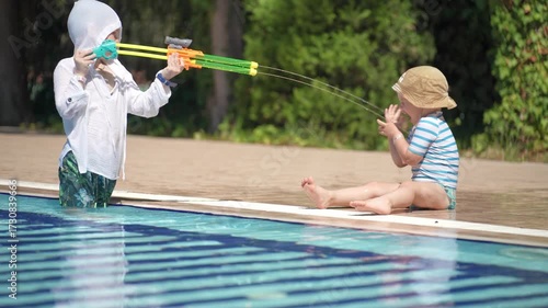 A happy little boy is enjoying himself in the pool. He is playing with a water gun and shooting at his younger brother.