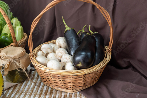 Fresh vegetables in a woven basket, featuring dark eggplants and white mushrooms, arranged on a textured surface with a rich brown backdrop, showcasing natural produce