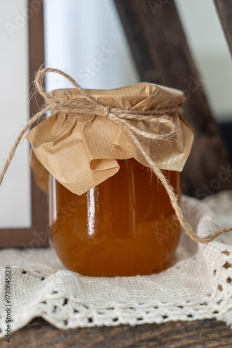 Glass jar filled with golden honey, topped with brown paper and twine, placed on a rustic wooden surface, surrounded by a delicate lace cloth, showcasing natural sweetness and warmth
