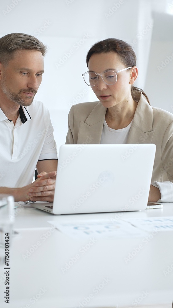 Fototapeta premium Businessman and adult businesswoman reviewing marketing analytics on laptops, working together strategically in contemporary office workplace. Business people concept