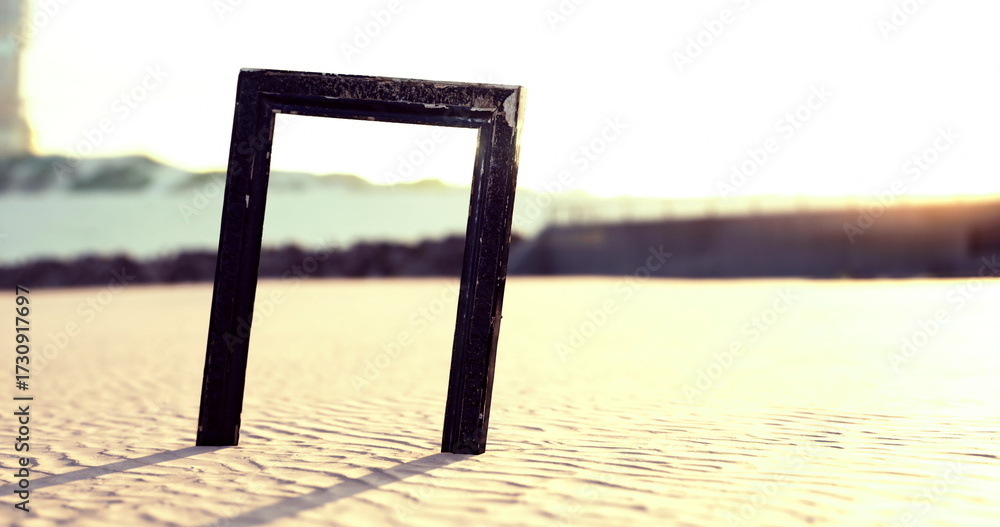 Fototapeta premium A black rectangular frame is positioned upright on a sandy beach during sunset. The warm light creates dramatic long shadows, enhancing the tranquil atmosphere of the seaside.