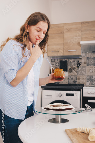 A beautiful young woman with long hair decorates s a chocolate sponge cake on a special stand an licks her finger, out of joy and delicious taste of salted caramel for glazing.