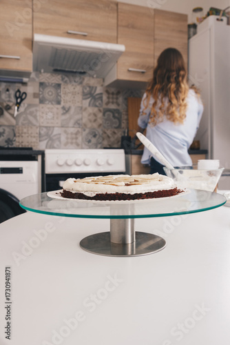 Young woman is cleaning on the kitchen and stays with hr back after preparation and decoration a chocolate cake with milk cream and bananas. Home made chocolate sponge cake, traditional cooking.