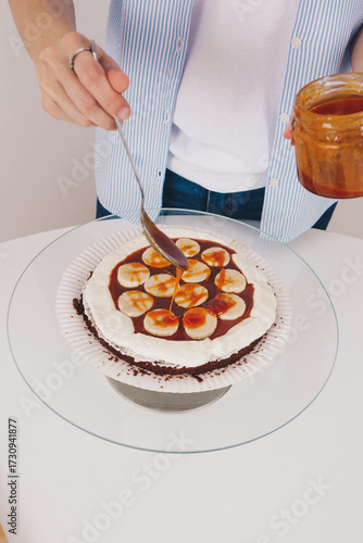 Young woman with slim hands putting salty caramel to a sponge cake on a special stand to assemble a delicious cake. Home made chocolate sponge cake, traditional cooking, young housewife.