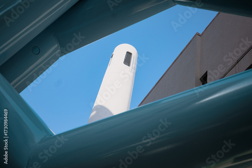 A creative architectural shot frames a white tower between glossy turquoise steel beams and a clear blue sky, showcasing the contrast of modern design with traditional elements.