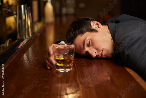 Man in suit lies forehead on bar beside whiskey glass, capturing inebriation and exhaustion in dimly lit pub scene.