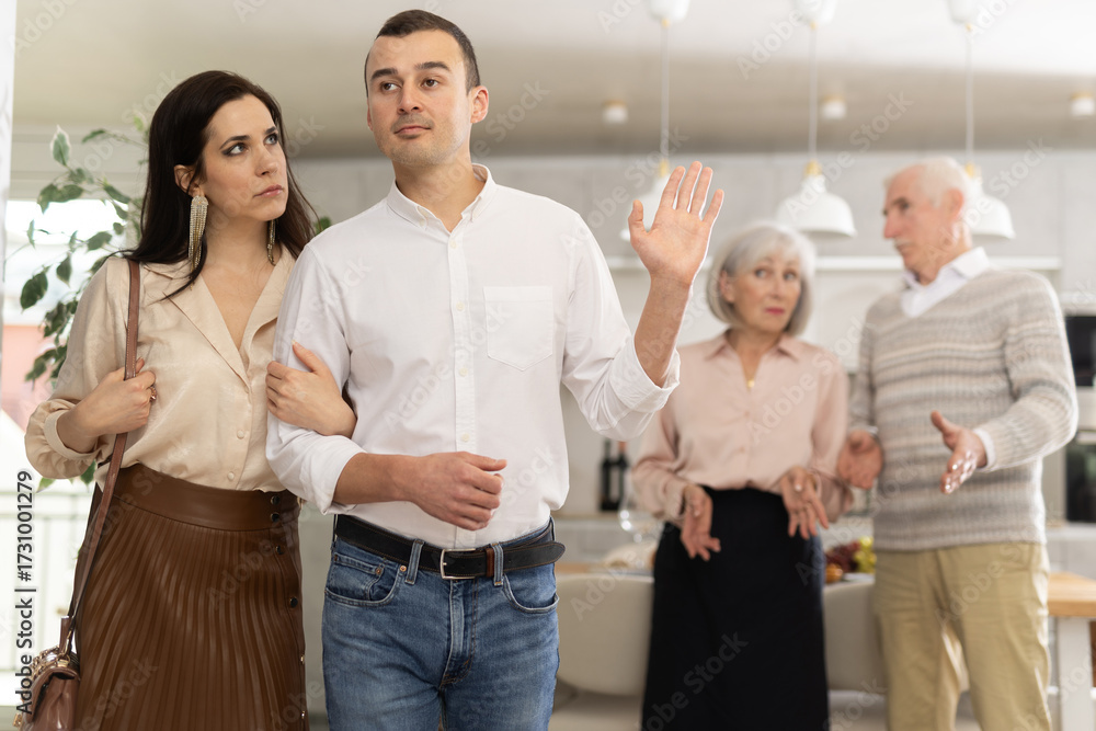 Obraz premium Sad young couple leaving the house while senior parents standing behind and looking at them discontentedly