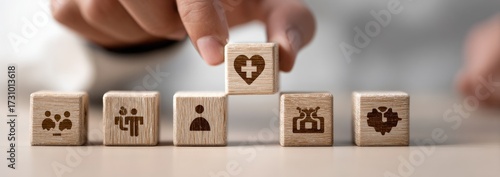 A hand places a wooden block with a heart and cross symbol on top of a stack of blocks with various icons