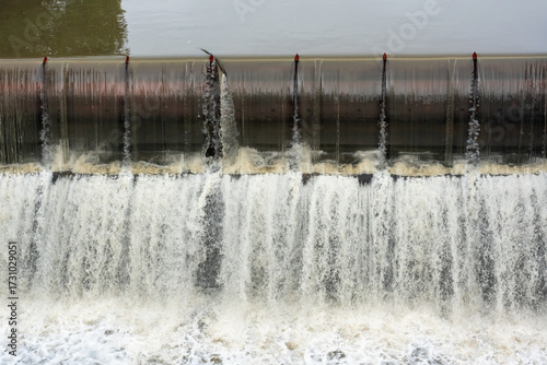 Concrete weir dam with multiple water jets and cascading waterfall creating white foam and spray. Urban water management infrastructure and hydraulic engineering structure in action.