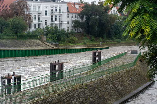 Flooded river with muddy turbulent water rushing past green flood barriers and white historic buildings. Concept for urban flood management and emergency response infrastructure systems.