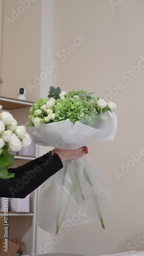A florist adjusts the white packaging on a white and green bouquet of roses, hydrangeas, and eucalyptus with his hands.