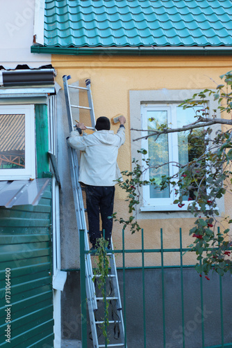 Man standing on a ladder painting the exterior wall of a house near the window. Outdoor renovation and maintenance work scene. Perfect for illustrating construction, repair, or DIY projects