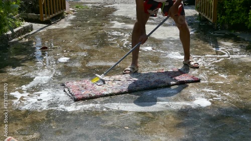 Close-up of a man washing a colorful carpet outdoors using a brush and soap, water flowing on the ground, traditional cleaning scene with summer light and strong shadows.