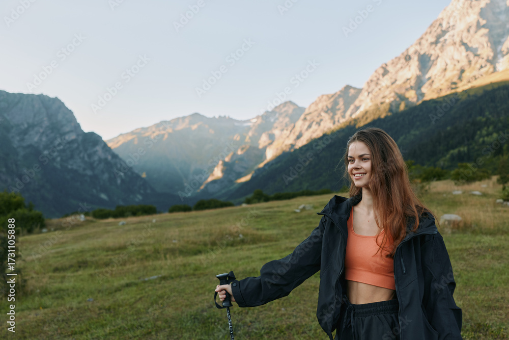 Fototapeta premium A cheerful woman hiker enjoys an outdoor moment in a mountain meadow. She smiles warmly, wearing a jacket and orange top, holding a trekking pole as sunlight highlights the alpine scenery.