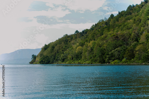 Wallpaper Mural Foto lanskap alam yang menampilkan keindahan danau dengan air biru jernih, dikelilingi perbukitan hijau yang rimbun. Di tepi danau terlihat rumah-rumah sederhana serta pepohonan tropis yang asri, menc Torontodigital.ca