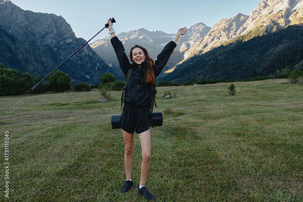 Fototapeta premium A cheerful hiker stands in a grassy valley with towering mountains in the background, raising a trekking pole and smiling, enjoying an adventurous outdoor moment.