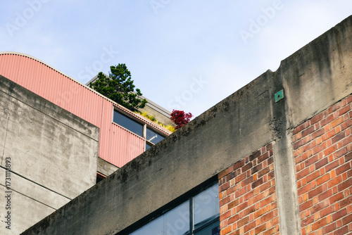 Low angle view of a concrete, red brick, and red-coated metal building exterior, with some balconies that have some plants just barely visible.