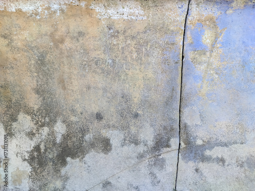 A weathered wall plaster, with a distinct vertical crack. The surface features a mottled texture with a blend of muted colors including beige, light blue, and various shades of gray and brown.