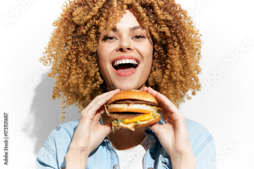 Joyful woman with curly hair holding a delicious burger, exuding happiness and enthusiasm against a plain background
