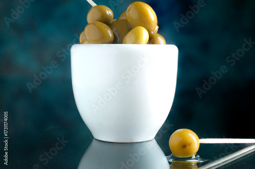 aceitunas dentro de un tazon blanco sobre una mesa de cristal y un fondo color azul, horizontal