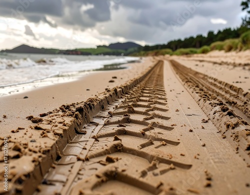 Tire tracks on a sandy beach recede toward a distant shoreline under a brooding sky