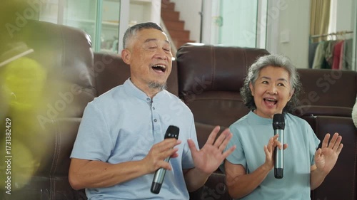 Asian elderly couple holding microphones and singing joyfully, celebrating anniversary with love and laughter at home