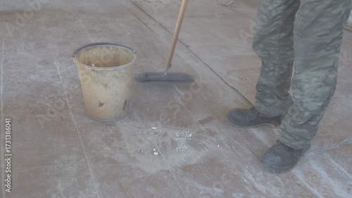 Man sweeping dust and debris on the floor of an empty, abandoned room using a broom or mop. The neglected space shows signs of decay and wear