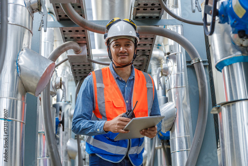 Fotografie Industrial engineer man inspecting equipment at factory plant, wearing safety helmet and vest, holding clipboard and Walkie Talkie, monitoring system performance in industrial environment