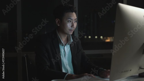 Asian businessman working late night at office desk in dark modern corporate environment with city lights in background, focused professional using desktop computer overtime after hours.