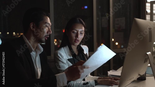 Businessman and businesswoman working late night in modern office, discussing project on printed documents, illuminated by computer screen, professional teamwork, deadline pressure, city lights.