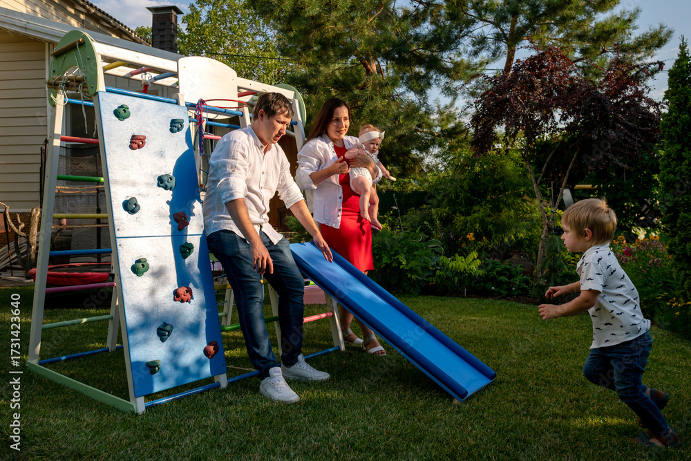 Fototapeta premium Parents crouch by a blue slide holding their baby as a toddler runs toward them, colorful playground set and green garden creating an active home scene