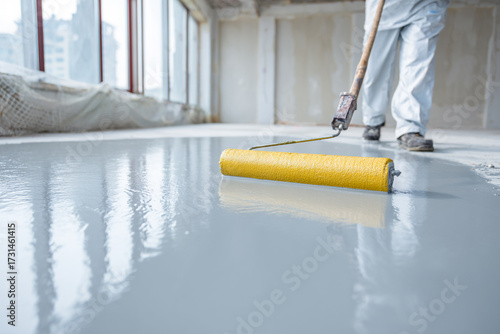 A construction worker in white protective gear applies a fresh coat of grey floor paint with a yellow roller