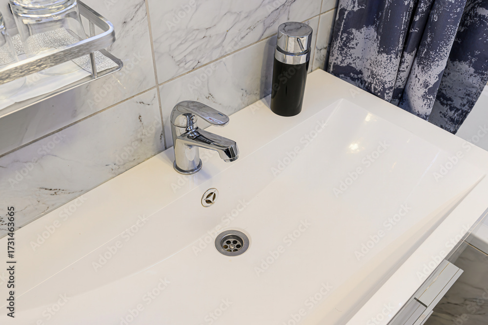 Fototapeta premium Close-up of modern white porcelain sink with chrome single-lever faucet and pop-up drain. Black matte soap dispenser, silver wire shelf holding glasses, gray marble-effect tiles