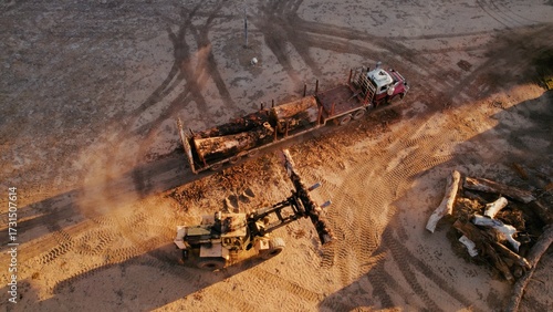 Heavy machinery operates on a construction site in a rural area at sunset, moving logs and preparing the landscape