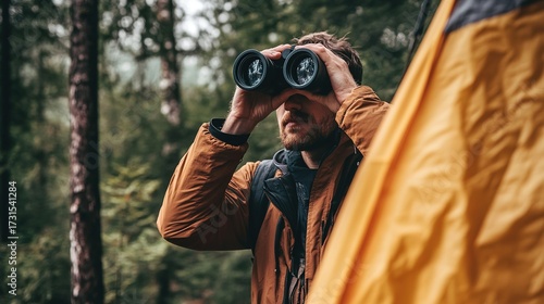 A man using binoculars while observing nature, standing near a tent in a lush forest environment.