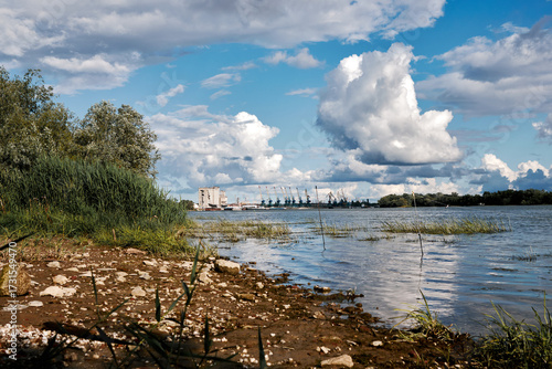 Scenic riverbank view with industrial cranes, cloudy sky, and natural shoreline