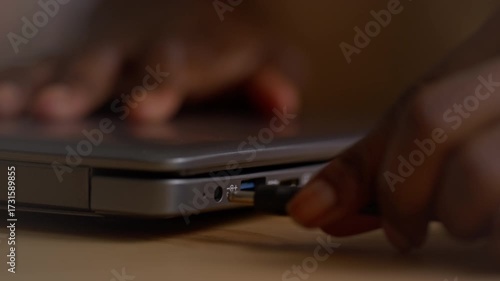 Selective focus close-up of hand of unrecognizable black woman plugging power cable into socket on laptop computer, while charging battery from electricity mains