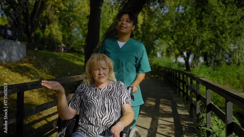Happy attractive senior female wheelchair user with age-related health conditions and black woman caregiver friendly communicating and sharing, enjoying leisure and morning stroll in summer park.
