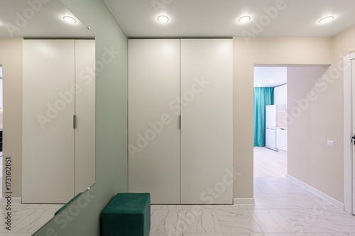 A modern apartment hallway with white marble floors, cream walls, and white storage cabinets. A green ottoman sits near a mirror