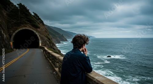 Man standing before road tunnel overlooking ocean on overcast day with rocky coastline and waves