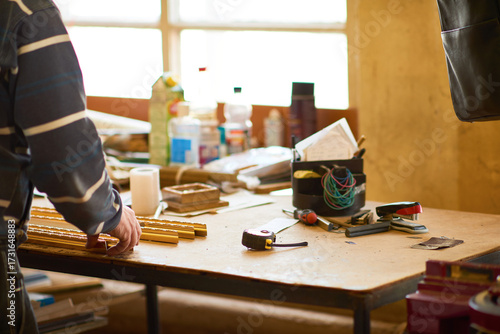 Man working with wooden picture frames on workshop table, using hands to arrange materials and tools, various supplies and equipment scattered in background