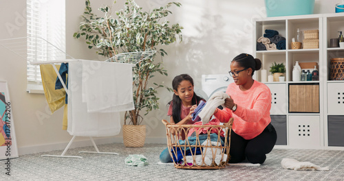 The daughter assists her mom in organizing laundry, folding colored clothes while standing near a dryer, with detergents and robes in the background.