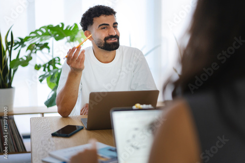 A young man with a beard and curly hair sits at a desk with a laptop, holding a pencil. He converses with a colleague, collaborating in a modern office environment.