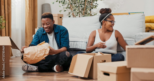 A couple enjoys the moment while sitting on the floor of their apartment. They carefully unroll items from boxes, discovering a photo frame, a keepsake vase, and meaningful trinkets.