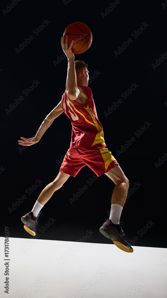 Obraz premium Basketball player in red and yellow uniform flying with ball above head in emotional action. Concept of sports branding, posters, education visuals, fitness lifestyle, and powerful design.