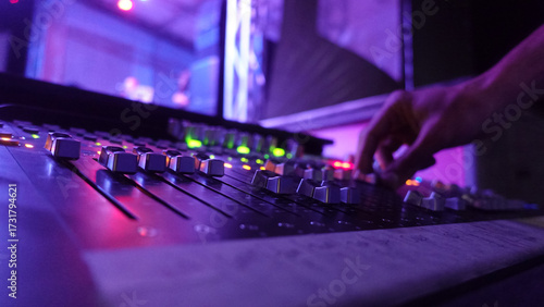 A close-up of a hand adjusting faders on a professional sound mixing console, illuminated by vibrant purple and pink stage lights. Perfect for music, live events, or audio production concepts.