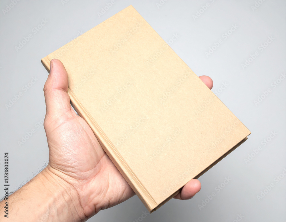 Obraz premium A person's hand holding a plain, unadorned brown book against a neutral gray background, highlighting the simple object