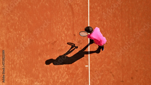 Tennis player in pink shirt practices on clay court, viewed from above. Flat lay photo highlights focused stance and shadow, emphasizing dedication and skill in sports training.