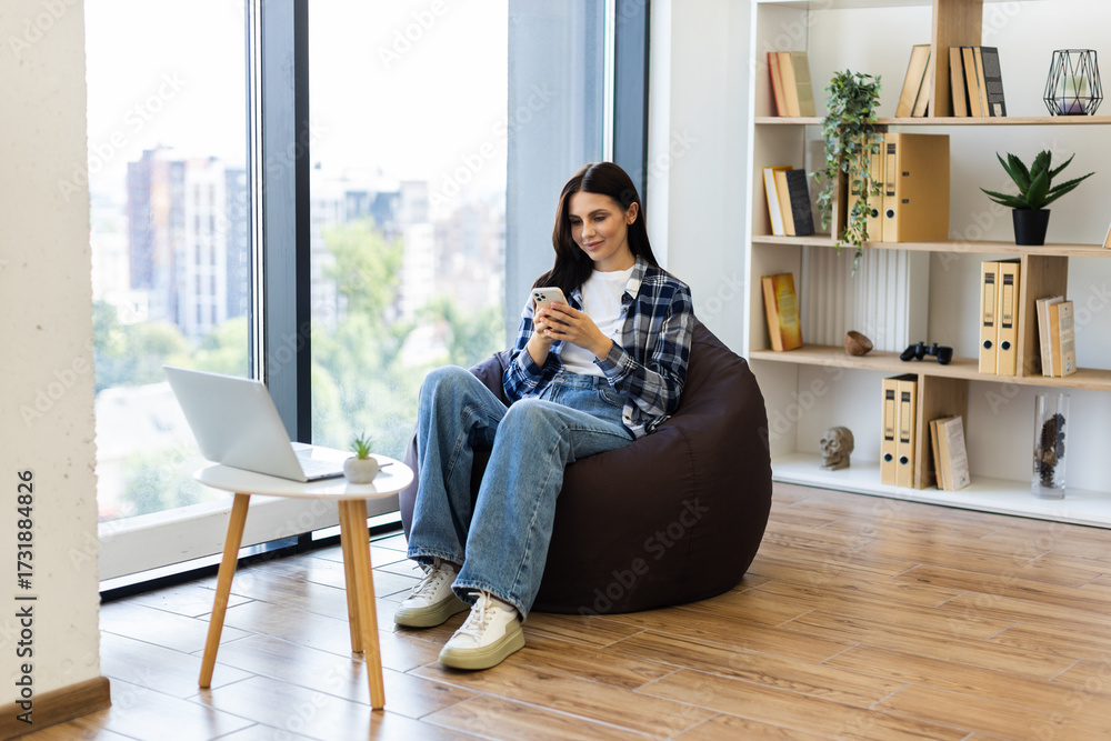 Obraz premium Young adult wearing casual clothes sitting on bean bag using smartphone near window. Interior features shelves with books and plants, wooden floor and bright daylight.